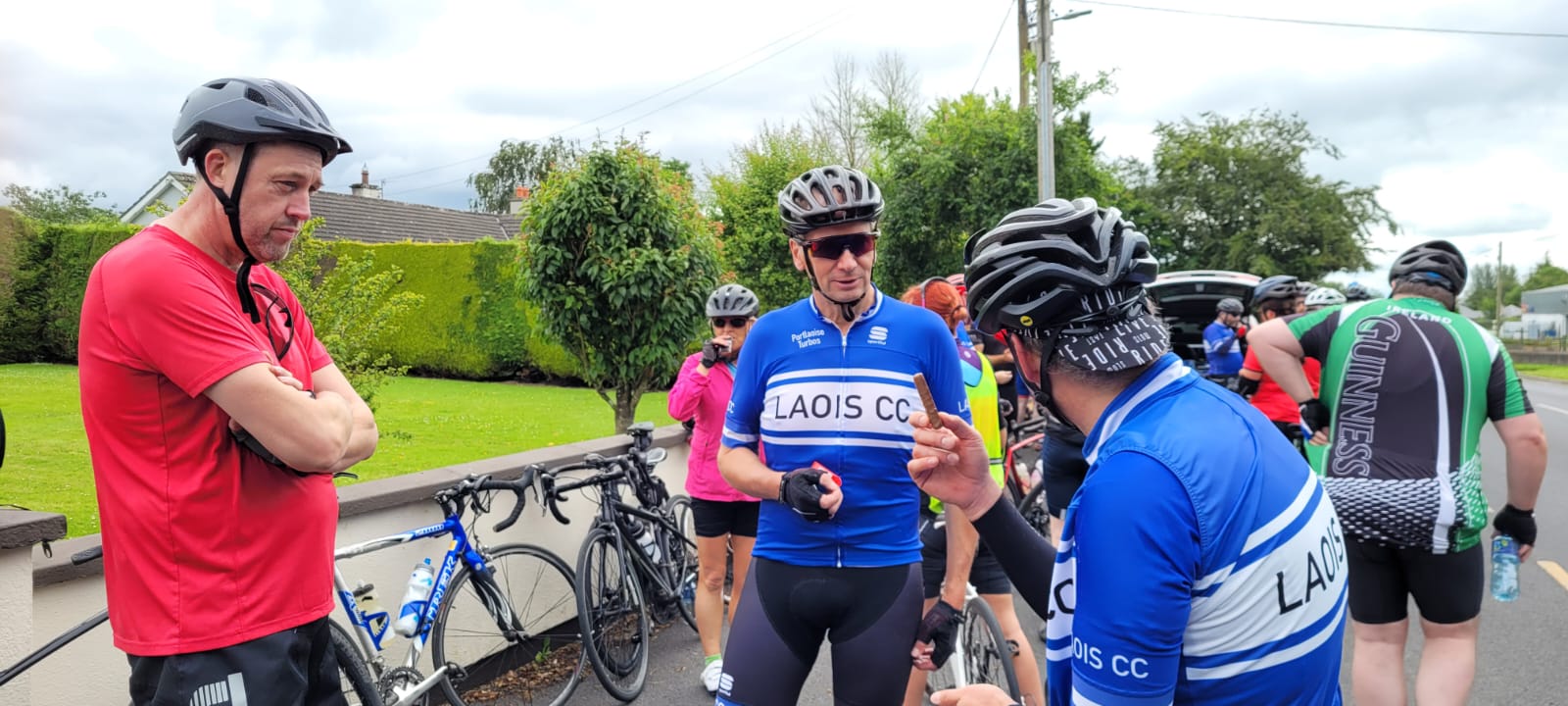 Three men wearing bike helmets speak with one another in bicycle gear at the side of a road.