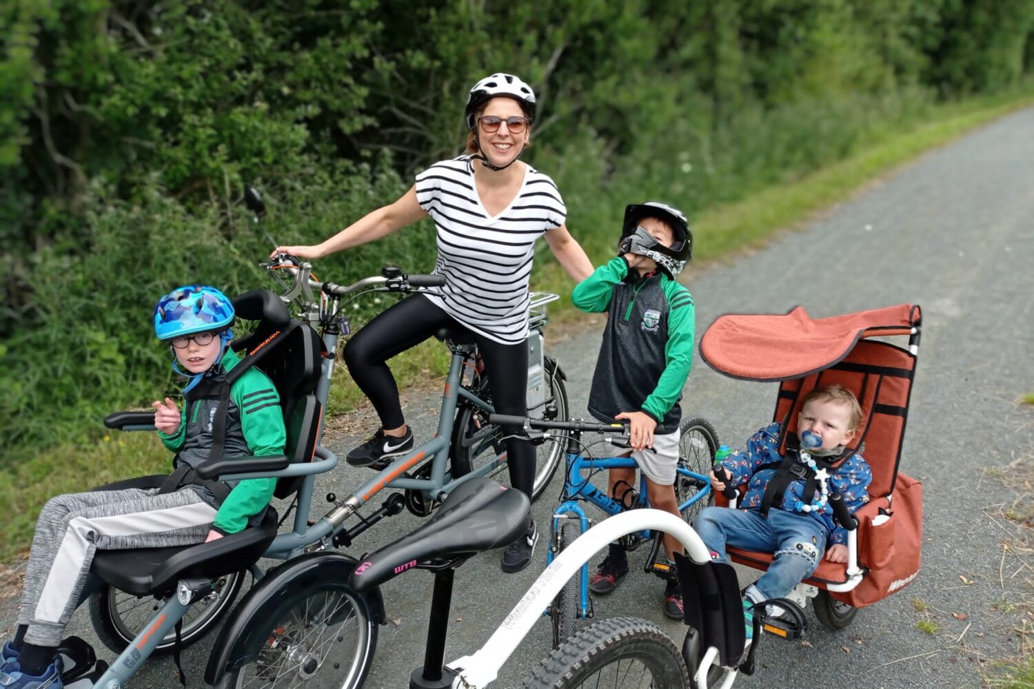 Mother cycles with three children in an accessible bicycle that can cycle with a wheelchair at its front.