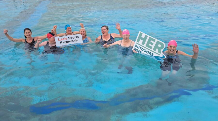 Group of women wave in pool holding signage for HER Outdoors week and Laois Sports Partnership.