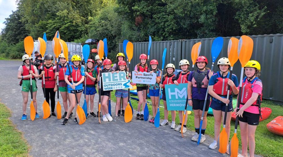 Group of young girls holding yellow and blue paddles. Standing with signage for HER Outdoors week and HER Moves.